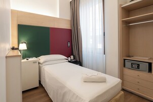 Habitación de hotel con cama, escritorio con lámpara, teléfono, mesita de noche con cafetera y armario.