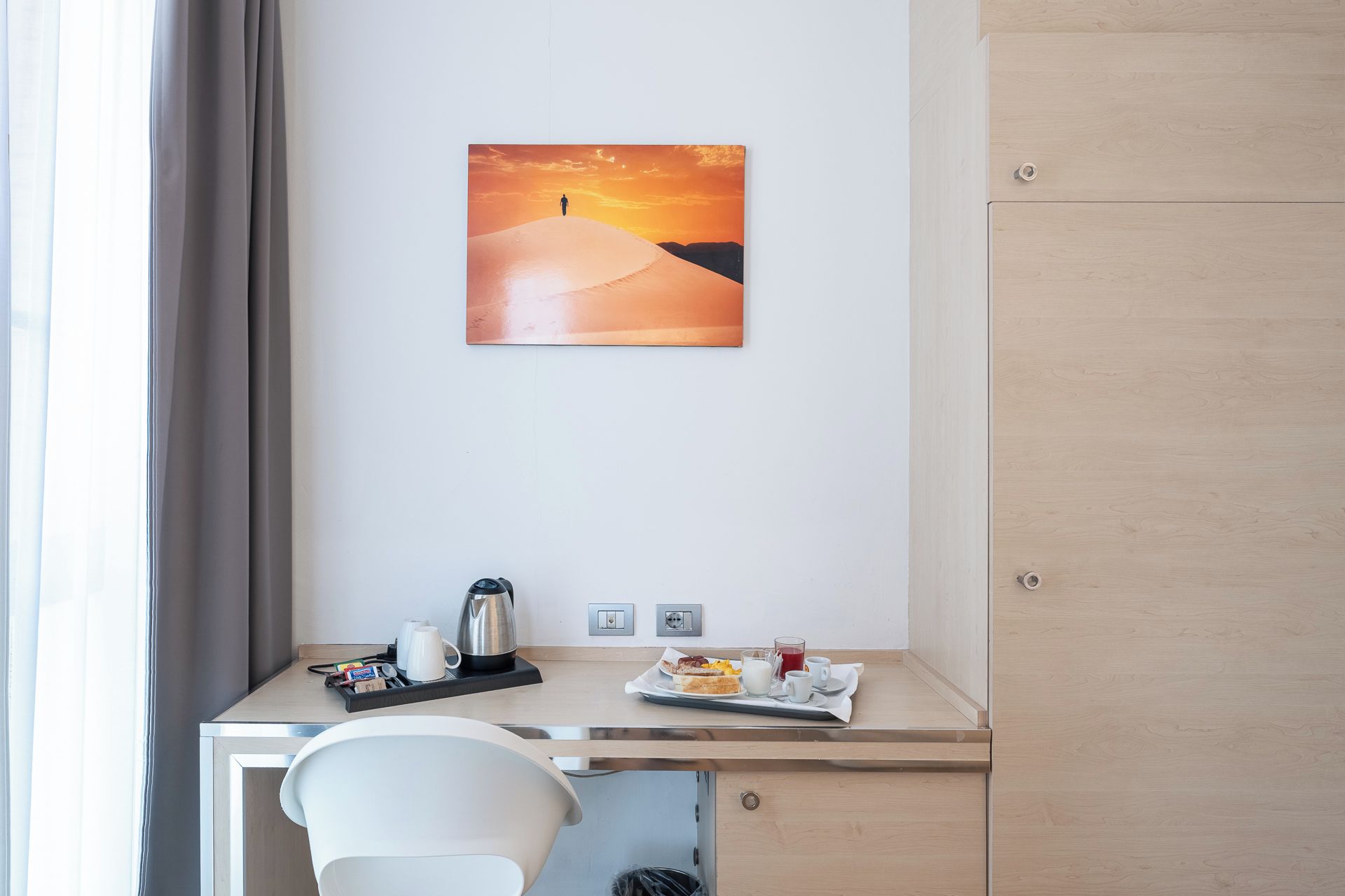 A hotel room with a desk, a chair, a painting of a desert landscape, and a breakfast tray with food and drinks.