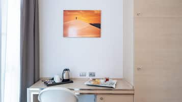 A hotel room with a desk, a chair, a painting of a desert landscape, and a breakfast tray with food and drinks.