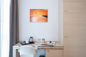 Habitación de hotel con escritorio, una silla, un cuadro de un paisaje desértico y una bandeja de desayuno con comida y bebidas.