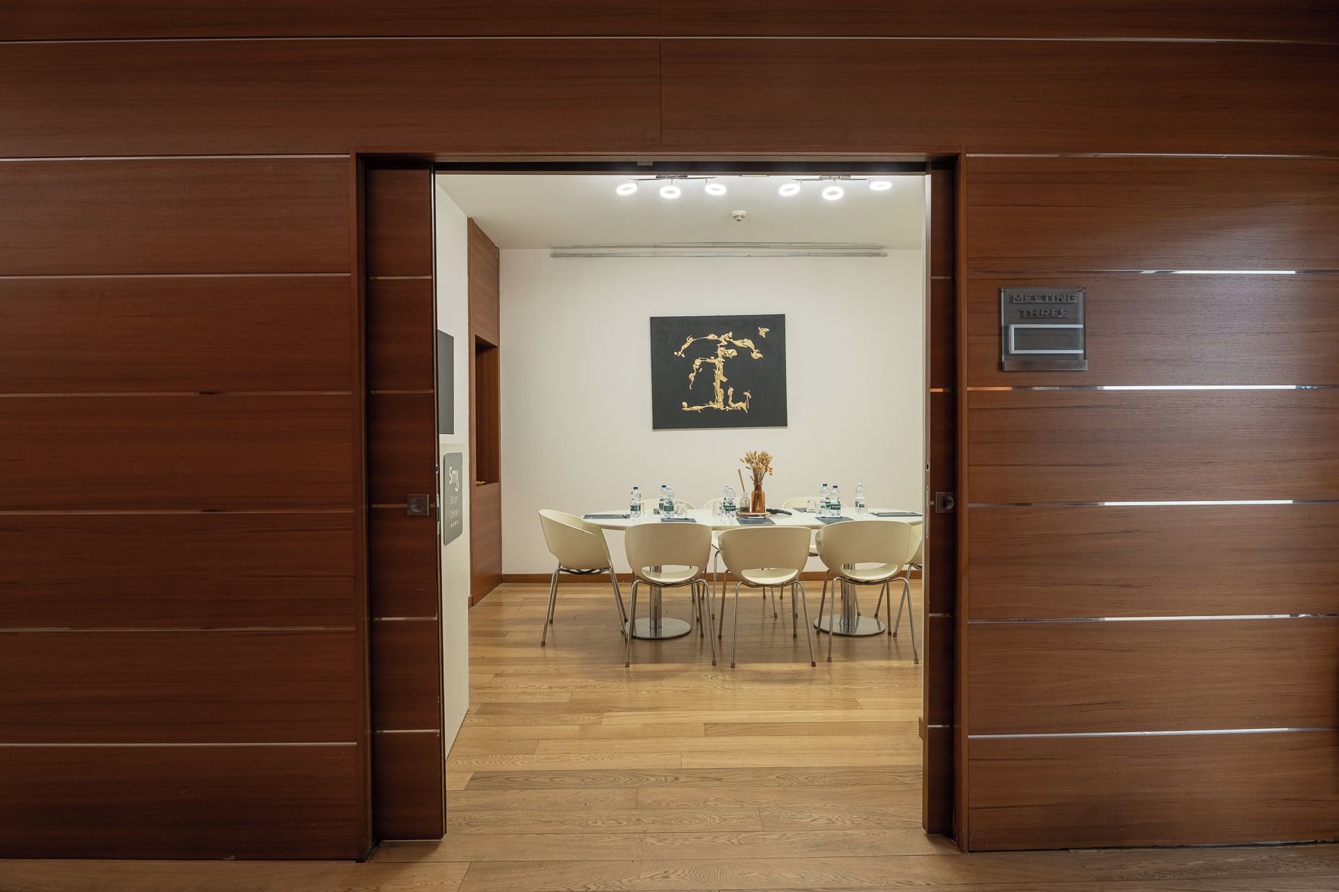 Een hotelkamer met een houten deur, een eettafel met stoelen, een aan de muur hangend kunstwerk en een bordje met het kamernummer.