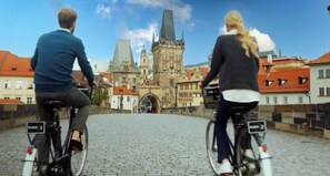 Dos personas andan en bicicleta por una calle adoquinada con edificios históricos al fondo.