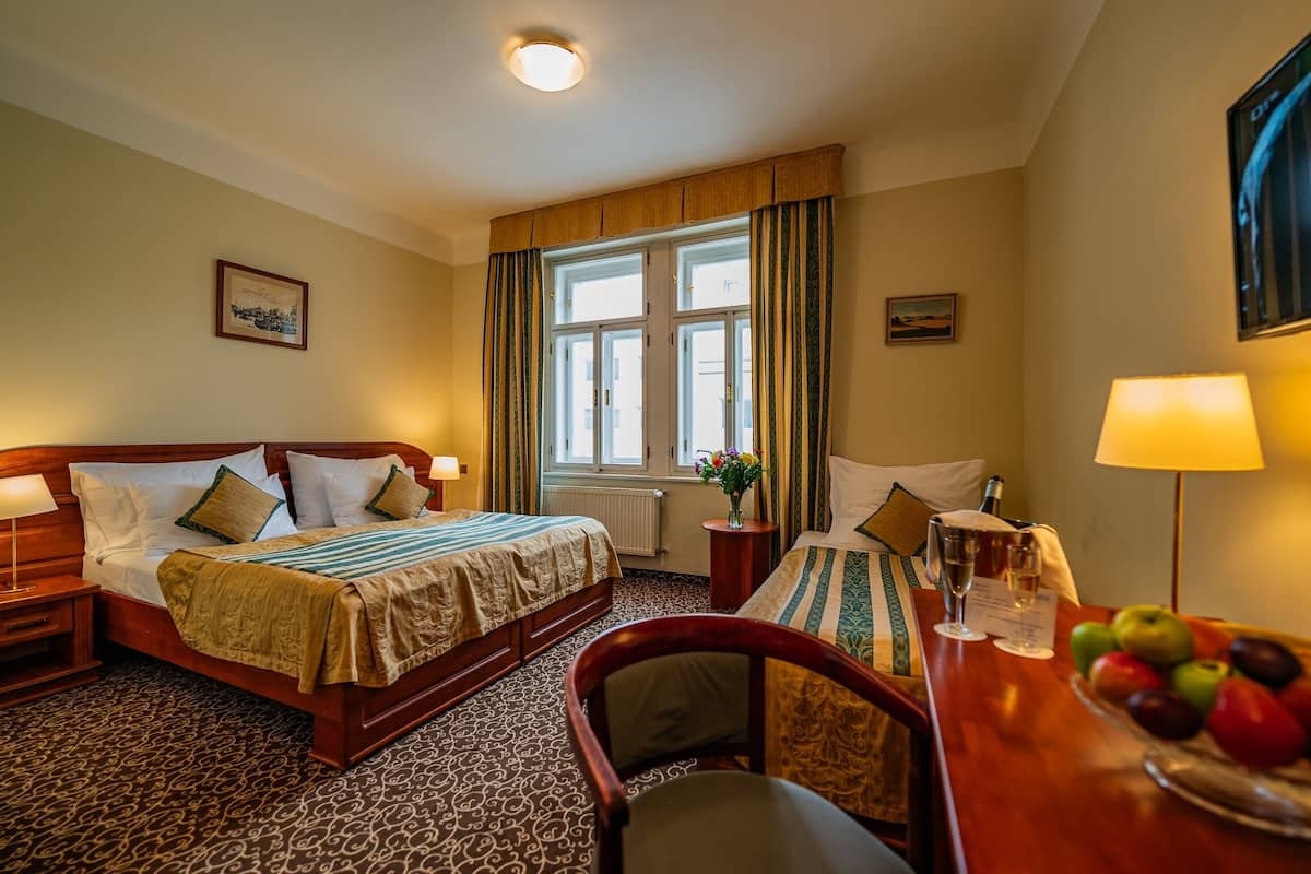 A hotel room with a large bed, a desk with a chair, a window with curtains, and a small table with a vase of flowers.