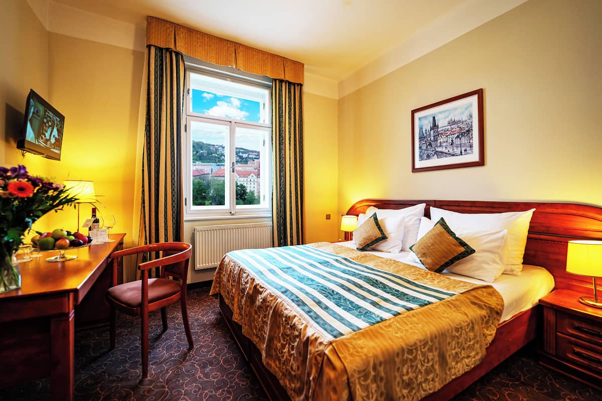 A hotel room with a large bed, a desk with a chair, a window with curtains, a framed picture on the wall, and a vase with flowers on the desk.