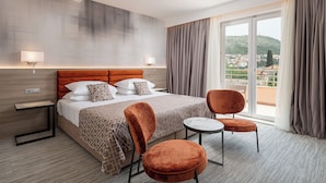 A hotel room with a large bed, two chairs, a small table, and a view of a cityscape through large windows. - Hotel Ivka (Dubrovnik)