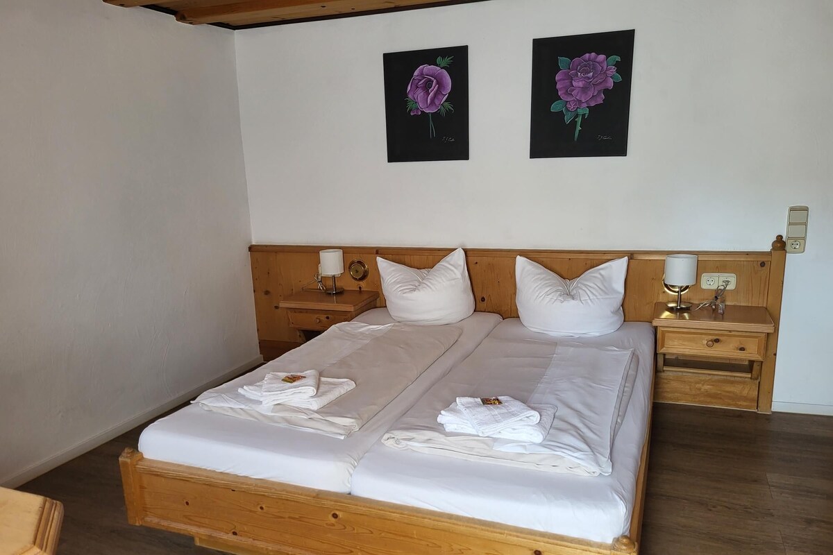 A double bed with white linens and towels, wooden headboard, two bedside tables with lamps, and two framed floral paintings on the wall.