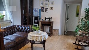 A living room with a brown leather sofa, a small table with a lace tablecloth, wooden flooring, a rocking chair, a water dispenser, and framed pictures on the wall. - Historikhotel Ochsen (Tamm)