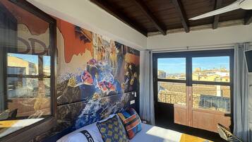 Una habitación de hotel con un ventanal grande, una cama con almohadas coloridas y un mural en la pared.