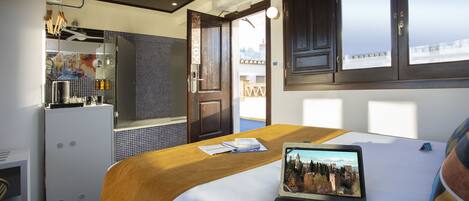 A hotel room with a bed, a view of a cityscape through the window, a small kitchenette, and a bathroom with a shower.