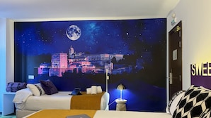 A hotel room with a large wall mural of a castle under a night sky, a bed with a laptop, a bedside table with a lamp, and a suitcase on the floor. - Casual Ilbira Granada Hotel by Casual Hoteles (Granada)