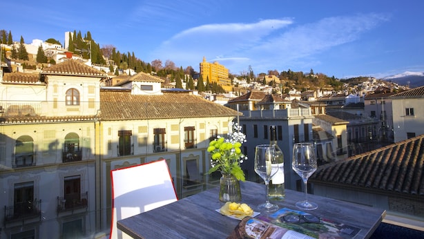 A table with a wine glass, a plate of chips, and a vase with flowers, overlooking a cityscape with historic buildings and a mountain in the background. - Casual Ilbira Granada Hotel by Casual Hoteles (Granada)