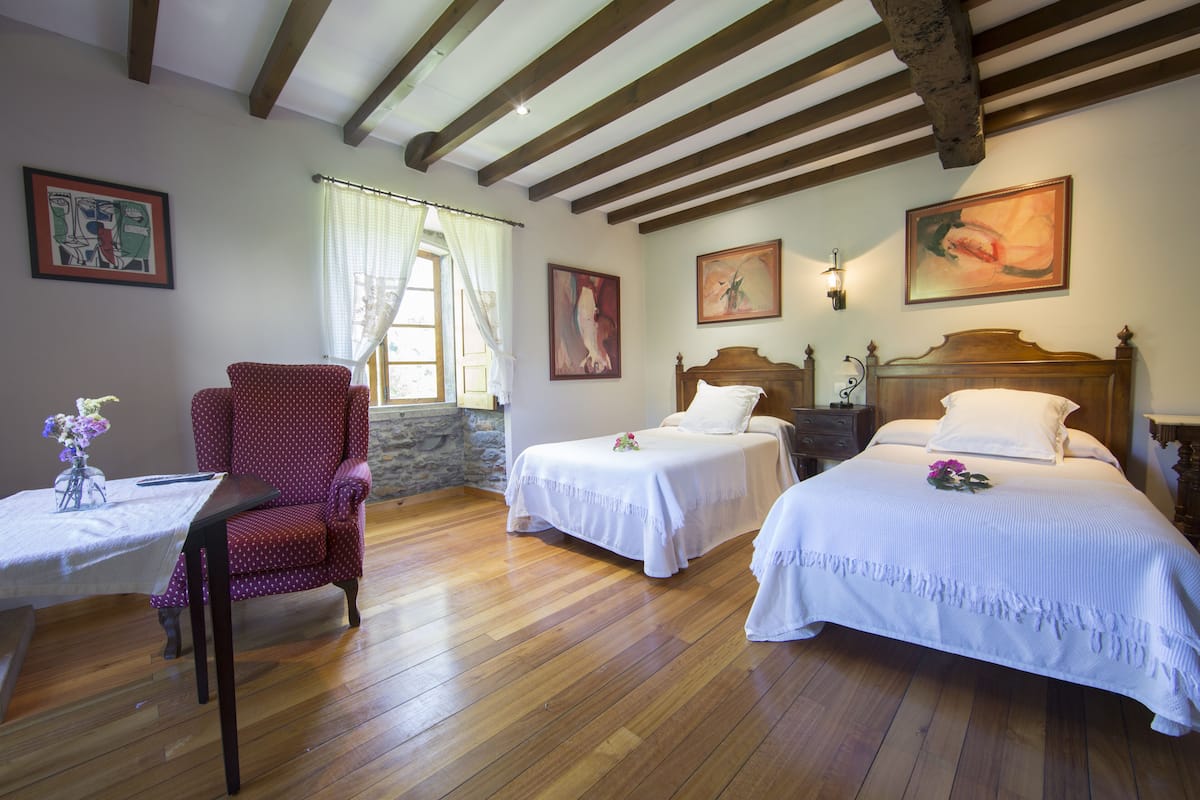 A room with two single beds, a wooden ceiling, a chair, and artwork on the walls.