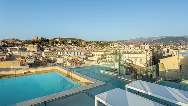 A rooftop pool area with clear views of a cityscape, including buildings and a hill with a castle-like structure.