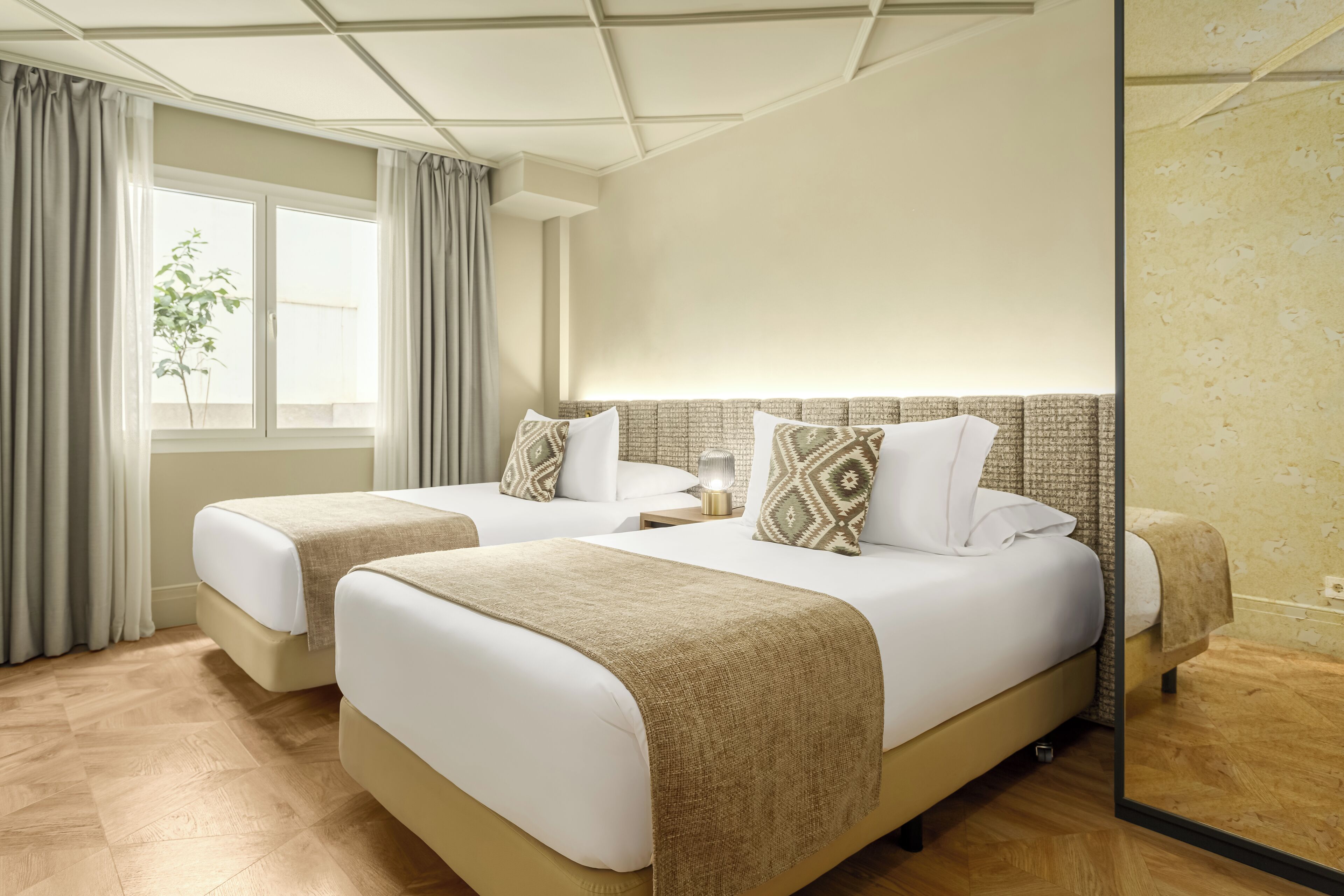 Una habitación de hotel con dos camas, un ventanal con cortinas, un espejo y suelo de madera.