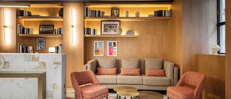 A modern lobby with a reception desk, a sofa, two patterned armchairs, a coffee table, and bookshelves with decorative items.