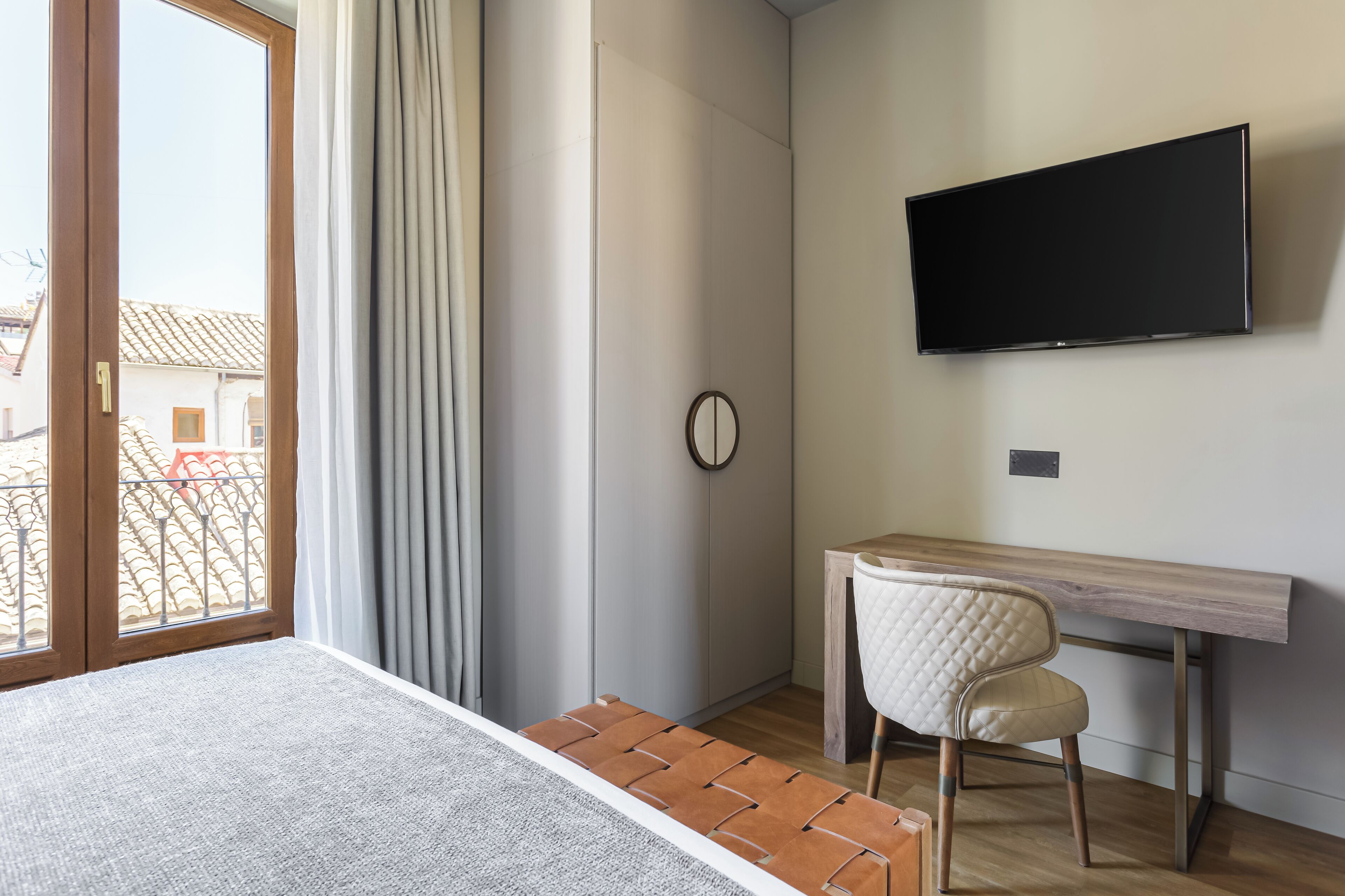 A modern room with a large window, a flat-screen TV mounted on the wall, a wooden desk, and a cushioned chair.