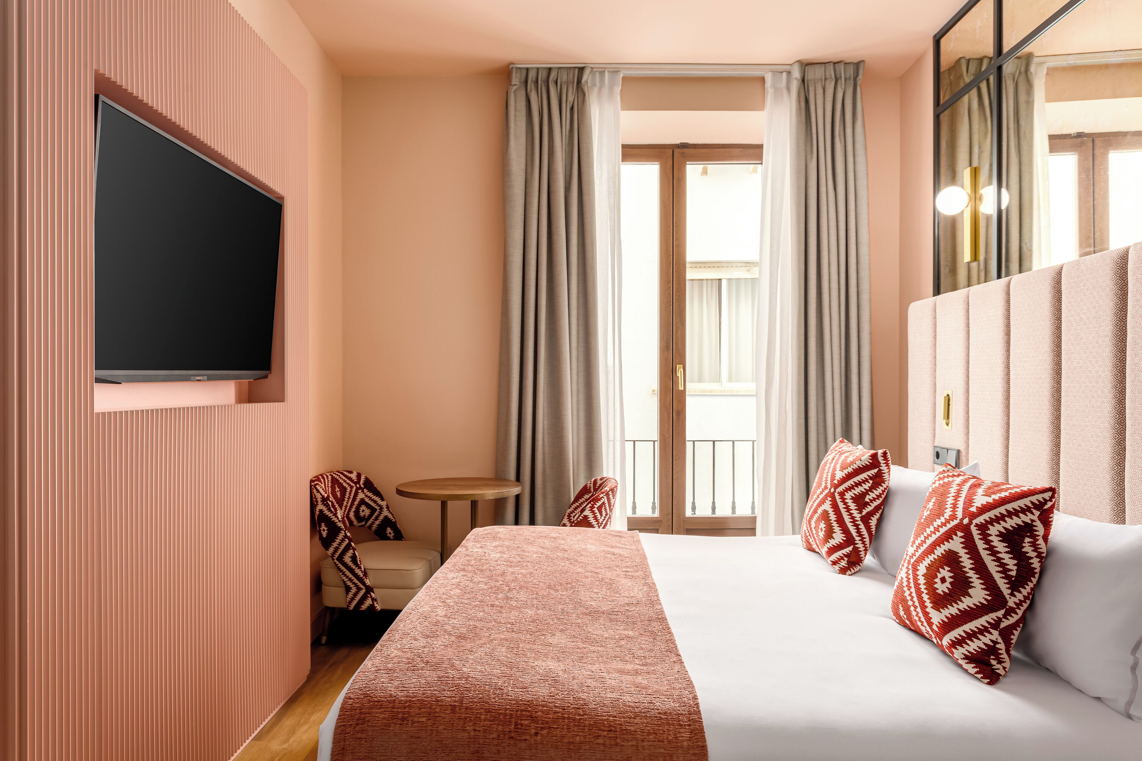 A hotel room with a large bed, a flat-screen TV mounted on the wall, a small round table, and a view of the outdoors through the window.