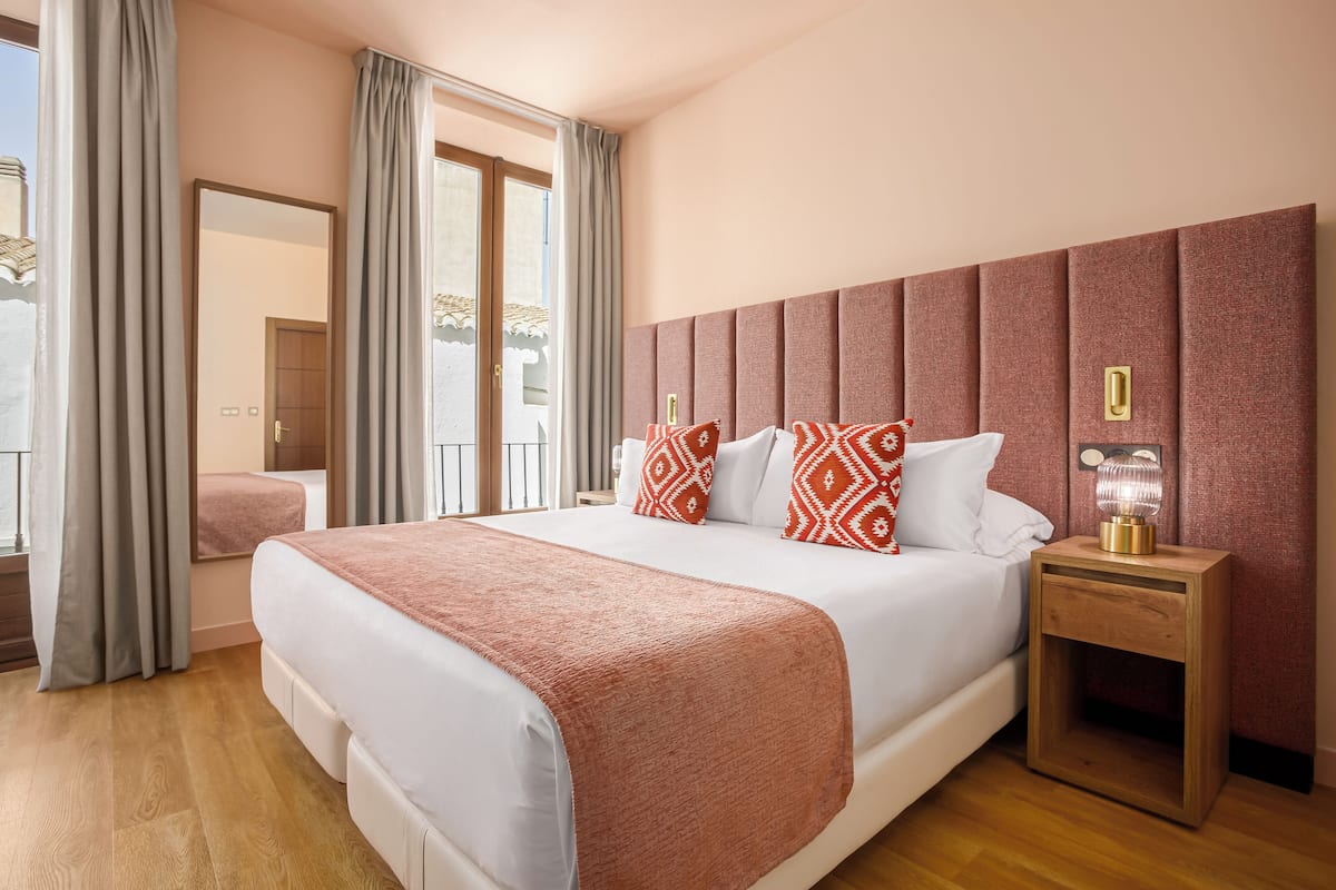 Ein Hotelzimmer mit einem großen Bett, einem Nachttisch aus Holz mit einer Lampe, einem Spiegel und Blick auf einen Balkon.