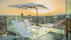 A glass-enclosed terrace with white lounge chairs and a large umbrella, offering a city view at sunset.
