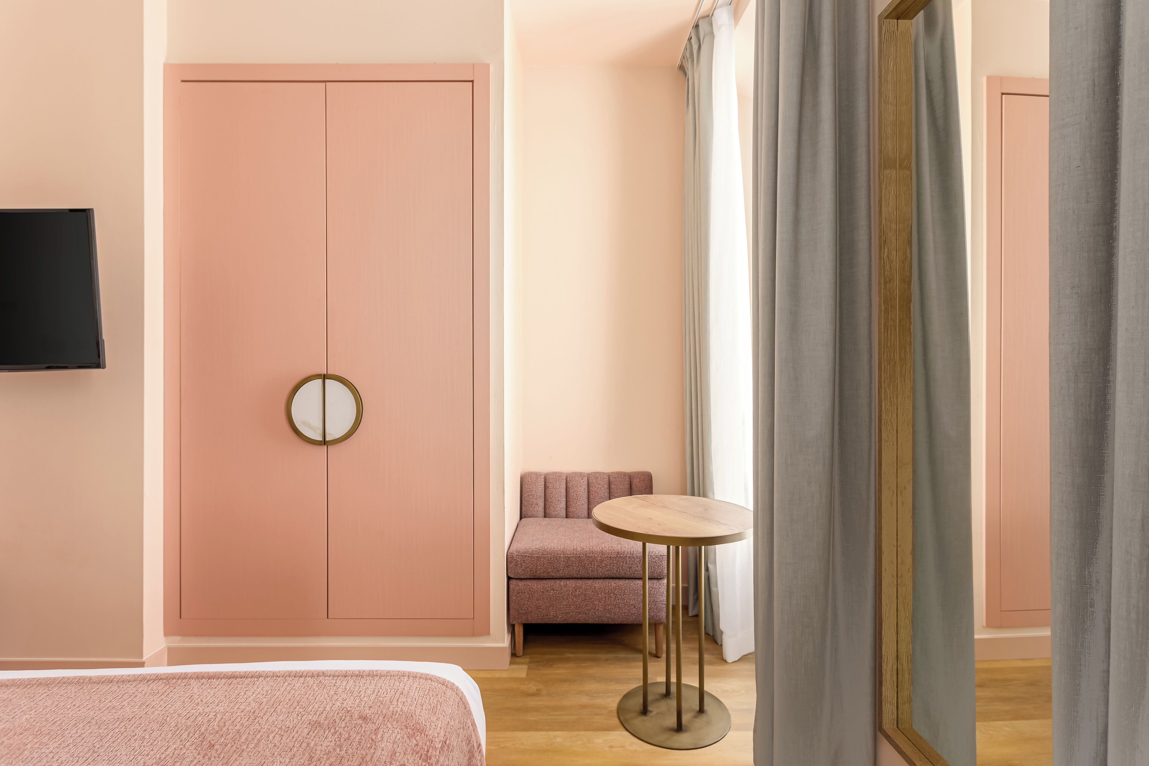 Una habitación de hotel con un armario rosa, una mesa redonda, un sofá y un televisor montado en la pared.