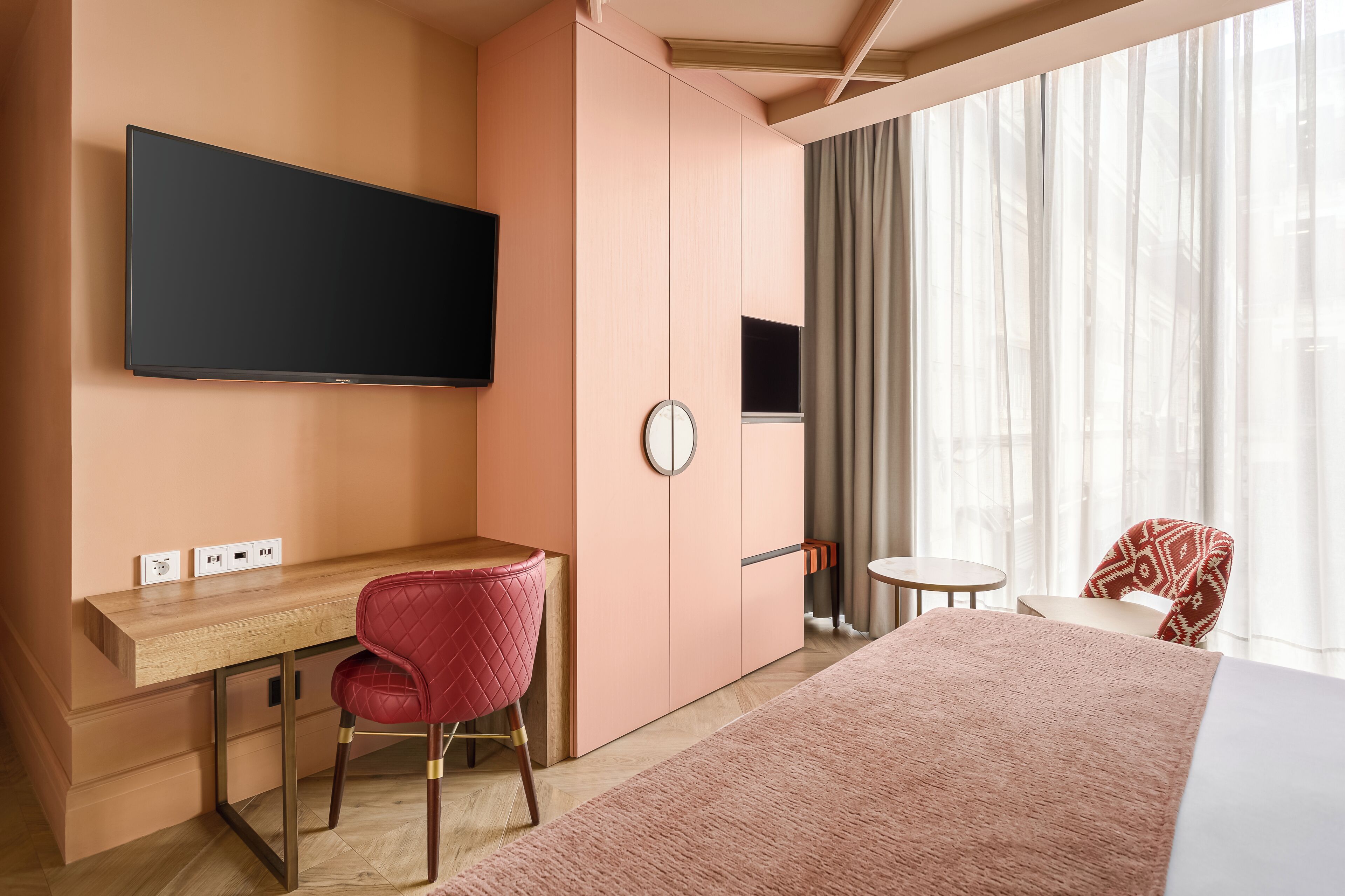 Una habitación de hotel con cama, escritorio con silla, televisor, reloj y ventana con cortinas.
