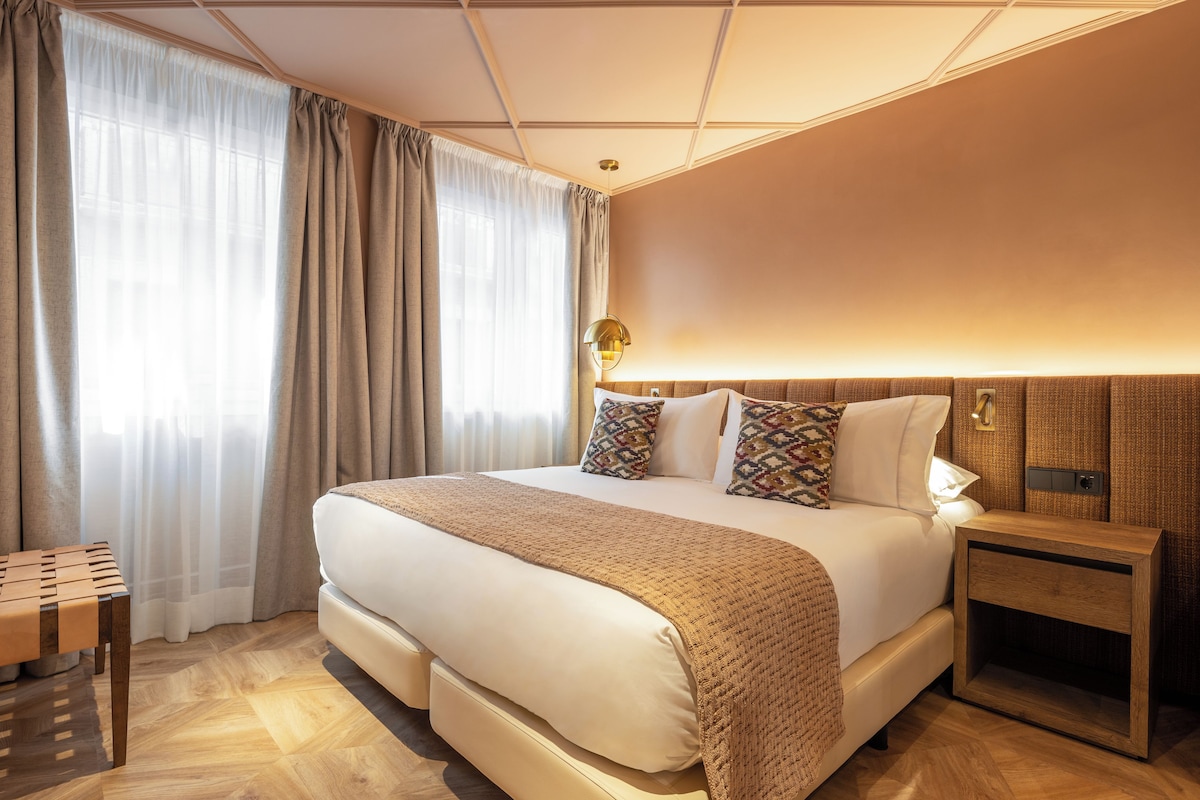A hotel room with a large bed, a bedside table, a lamp, and a window with curtains.