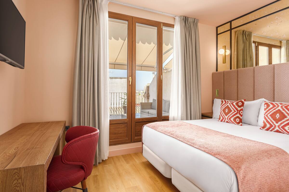 Ein Hotelzimmer mit einem Holztisch, einem roten Sessel, einem großen Bett mit gemusterten Kissen und einer Glastür, die auf einen Balkon im Freien führt.