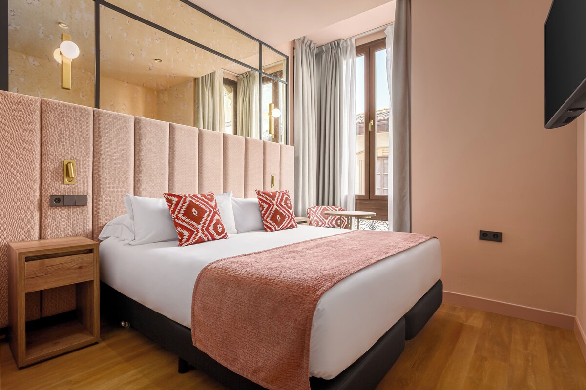 A modern hotel room with a large bed, a bedside table, a mirror, and a window with curtains.