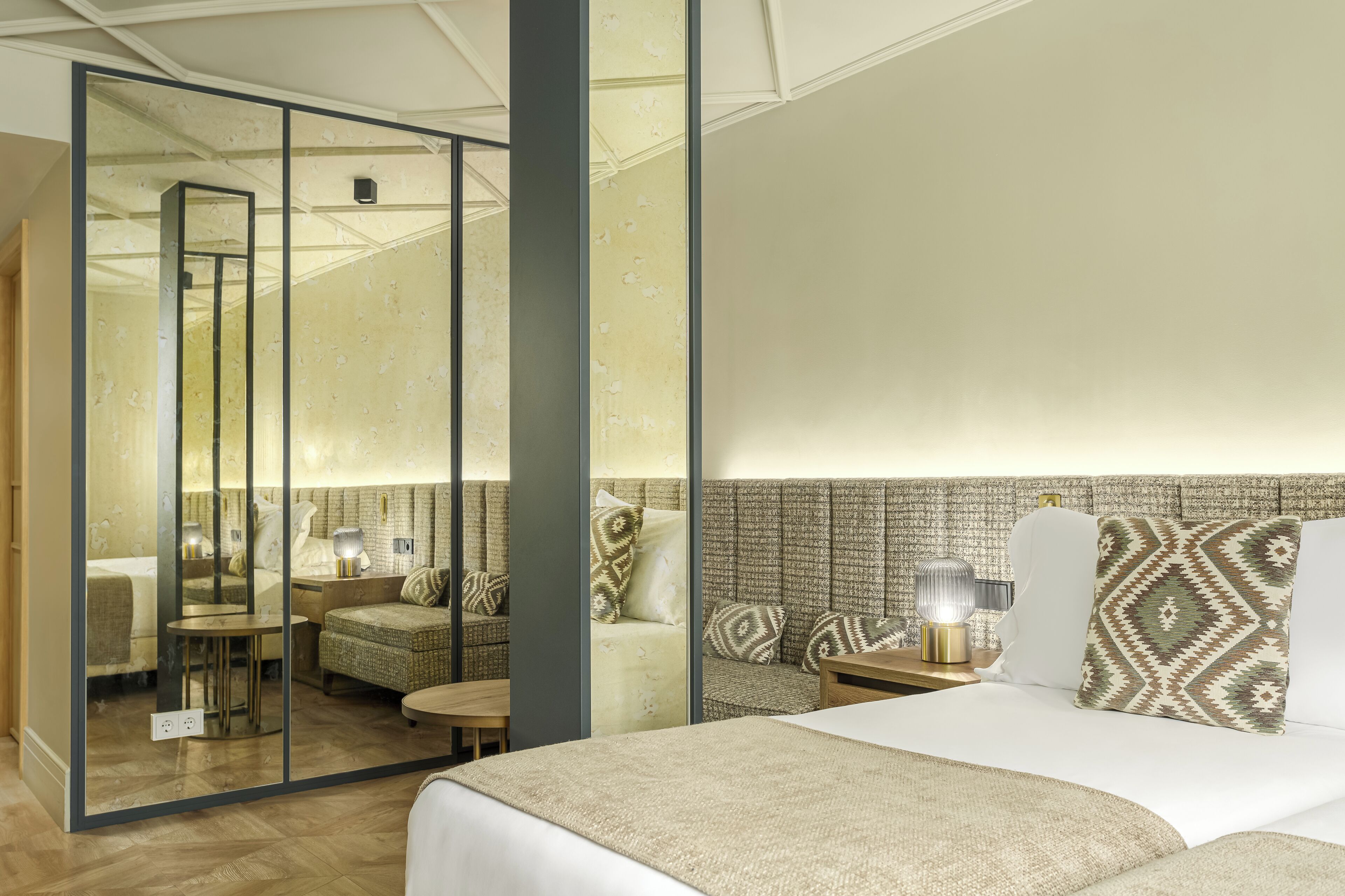 Una camera d'albergo moderna con un letto grande, un'area salotto con divano e poltrone, un tavolino e un pannello di vetro che divide l'ambiente.