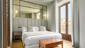 A hotel room with a large bed, a wooden bench, a nightstand, a mirror, and a view of the outside.