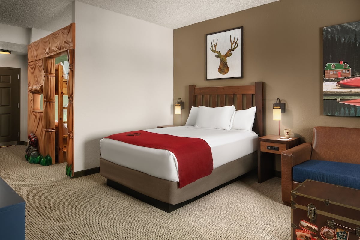 A hotel room with a large bed, a wooden headboard, a red throw blanket, a nightstand with a lamp, a suitcase, and a painting of a deer on the wall.