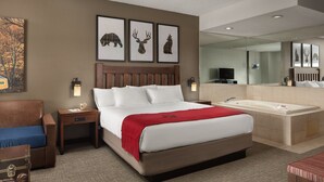Une chambre d’hôtel avec un grand lit, une tête de lit en bois, un plaid rouge, une table de chevet avec une lampe, une petite table avec une tasse, une valise, un grand miroir et une baignoire.