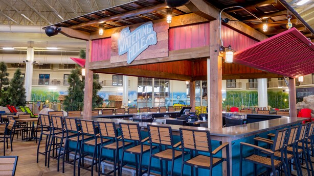 Un espace bar agrémenté d’une enseigne en bois sur laquelle on peut lire « The Watering Hole », avec un comptoir et des sièges comprenant des chaises et des tables.