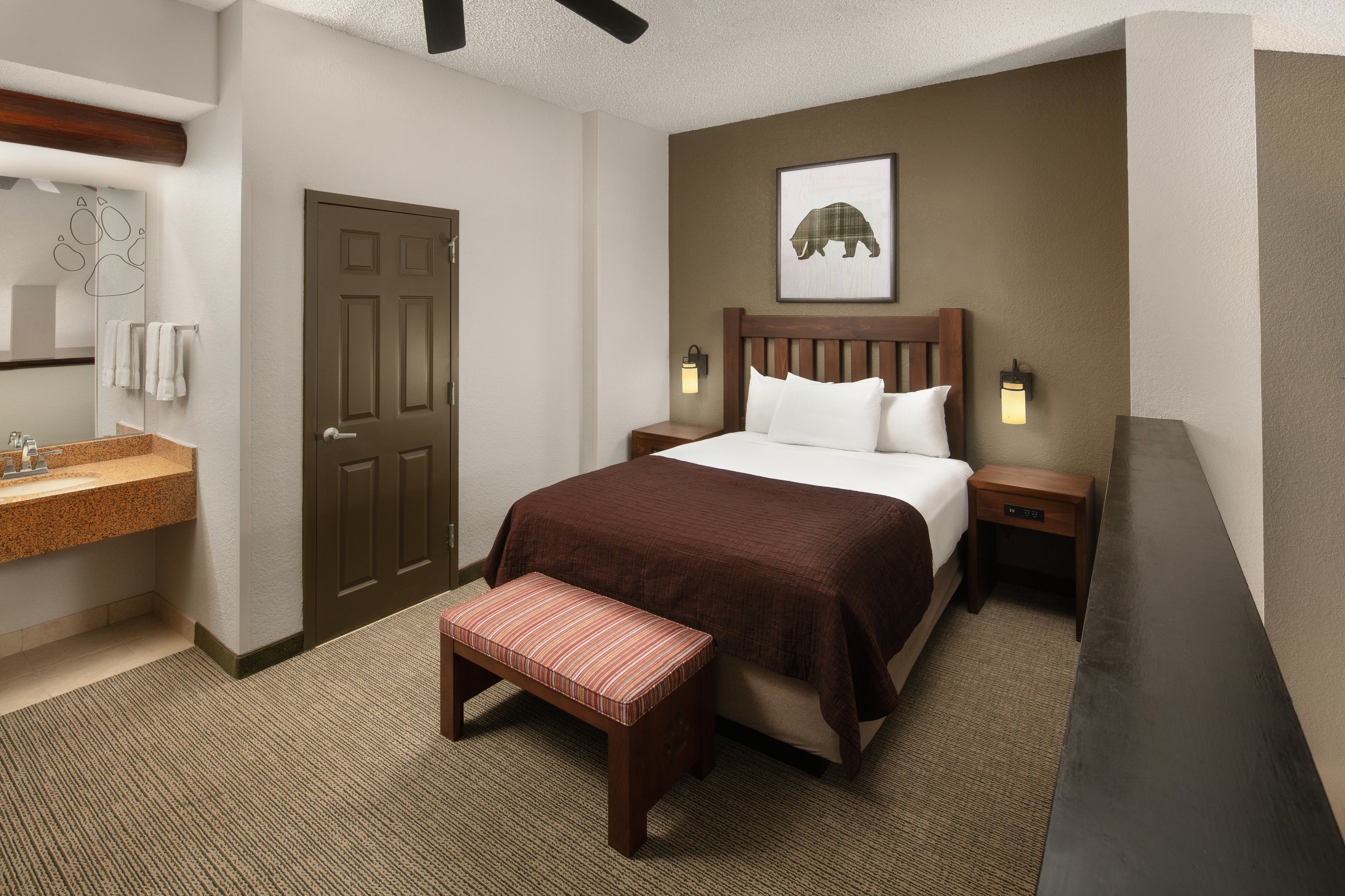 A hotel room with a bed, a bench, a nightstand, a wall-mounted light, a door, and a bathroom with a sink and towel rack.