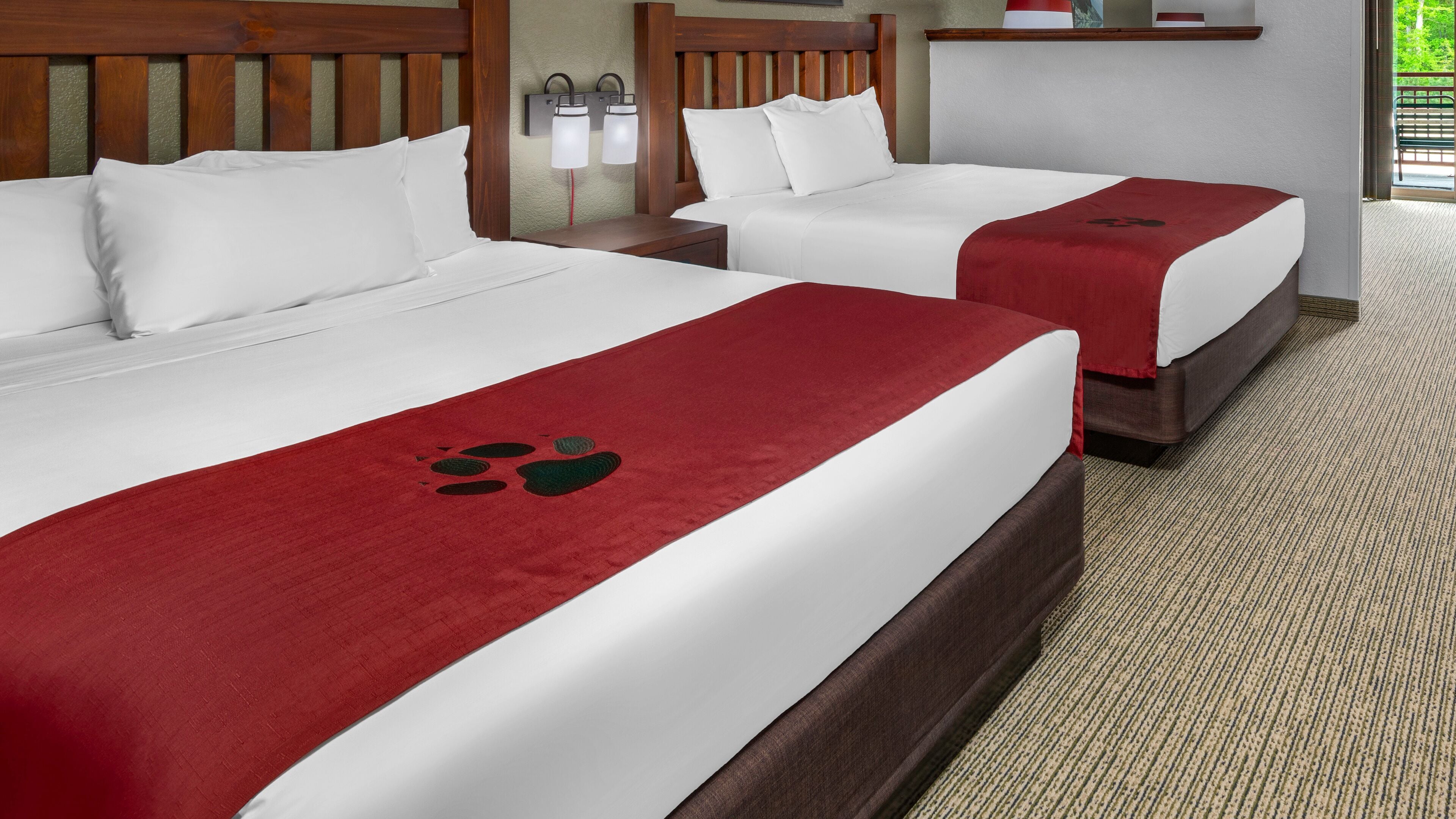 Two double beds with white linens and red bedspreads, wooden headboards, and a nightstand with a lamp.