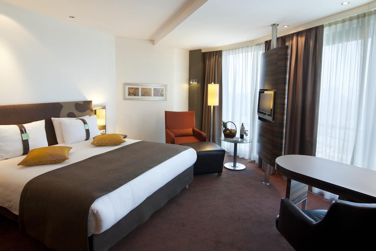 A hotel room with a large bed, a desk, a chair, a TV, and a small table with a lamp and fruit basket.