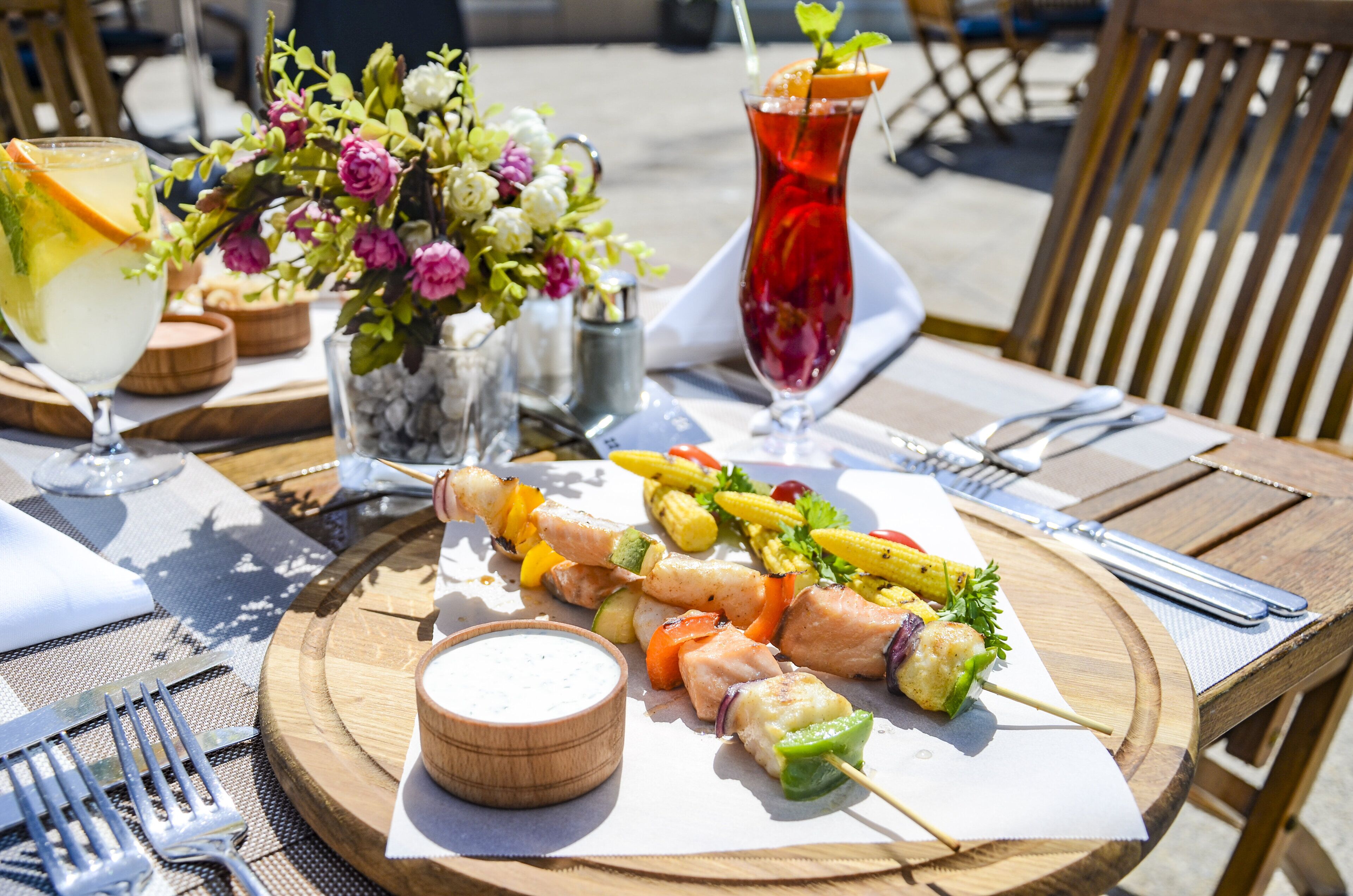A wooden table with a meal of grilled chicken skewers, corn, and vegetables, a drink with a slice of orange, and a small bowl of dipping sauce.