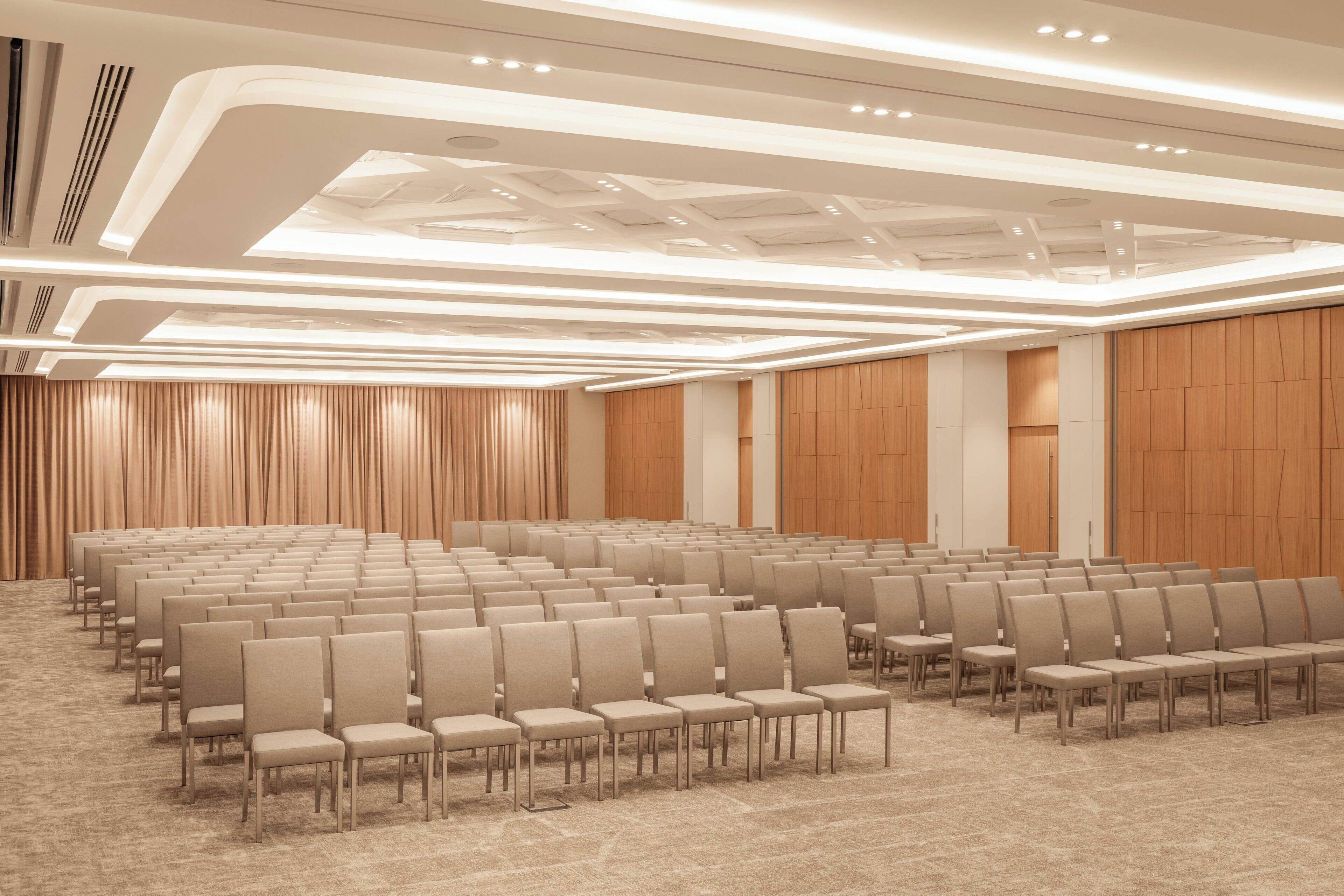 Un salón de conferencias con hileras de sillas, piso alfombrado y un diseño moderno en el techo.