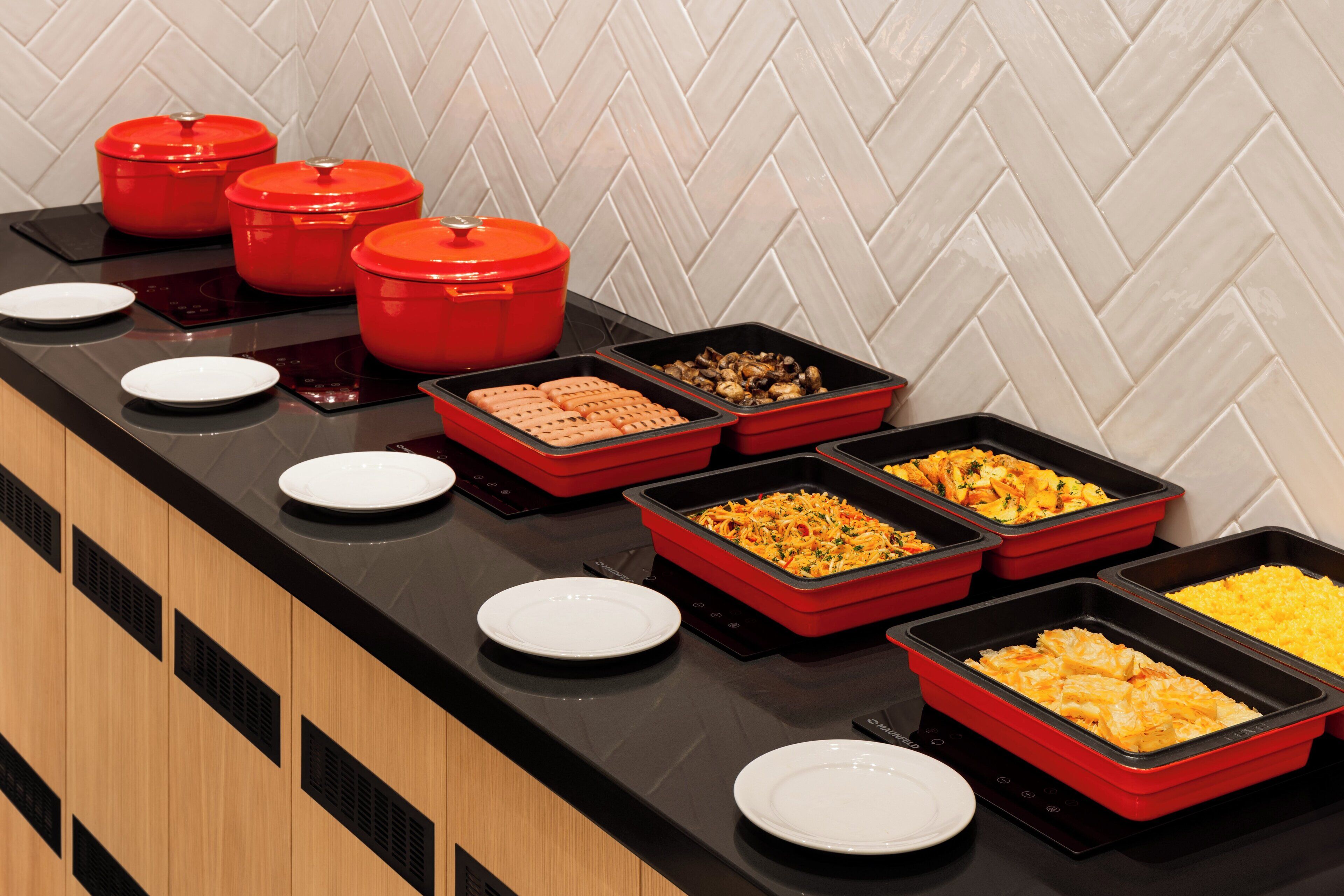 A buffet setup with various food items in red and black trays, accompanied by white plates.