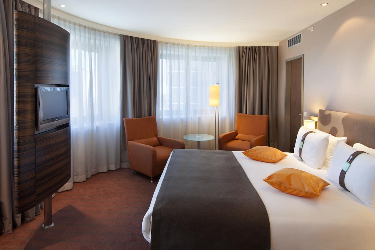 A hotel room with a bed, two armchairs, a TV, a lamp, and a view of the outdoors through large windows.