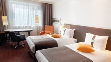 A hotel room with two beds, a desk, a chair, and a lamp.