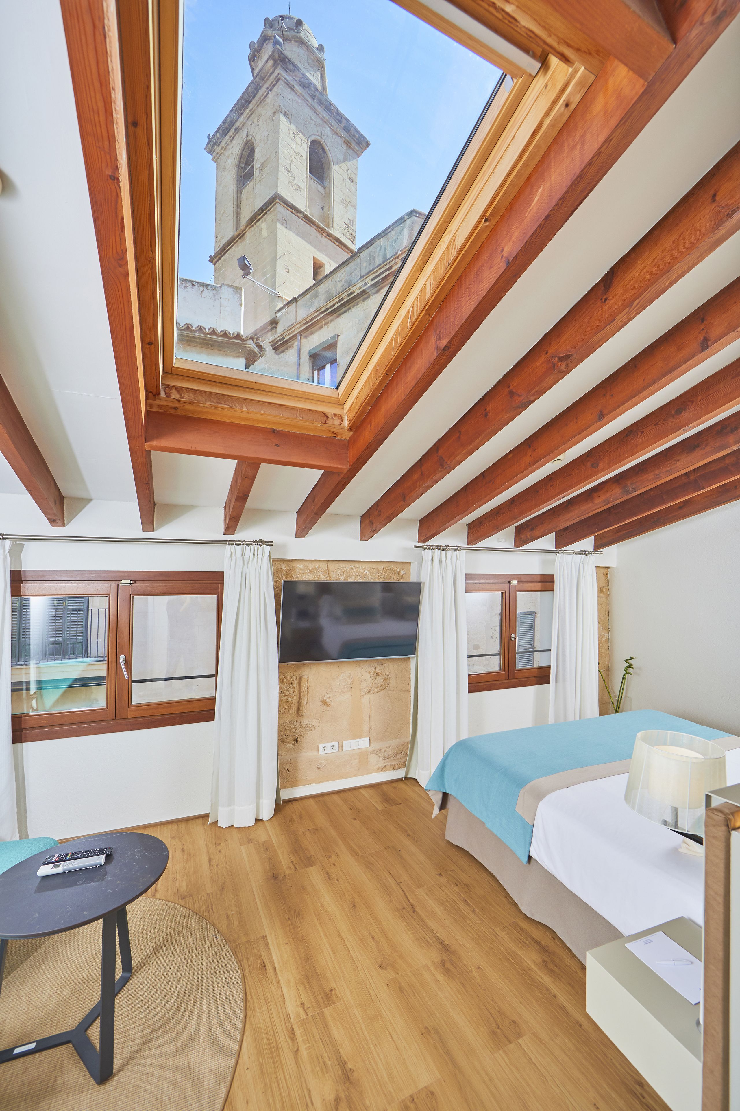 A room with a skylight, wooden beams, a bed, a table, and a view of a tower.