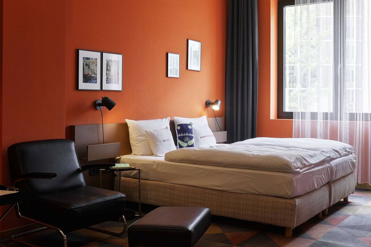 Quarto de hotel com cama, uma cadeira, escrivaninha e janela com cortinas.
