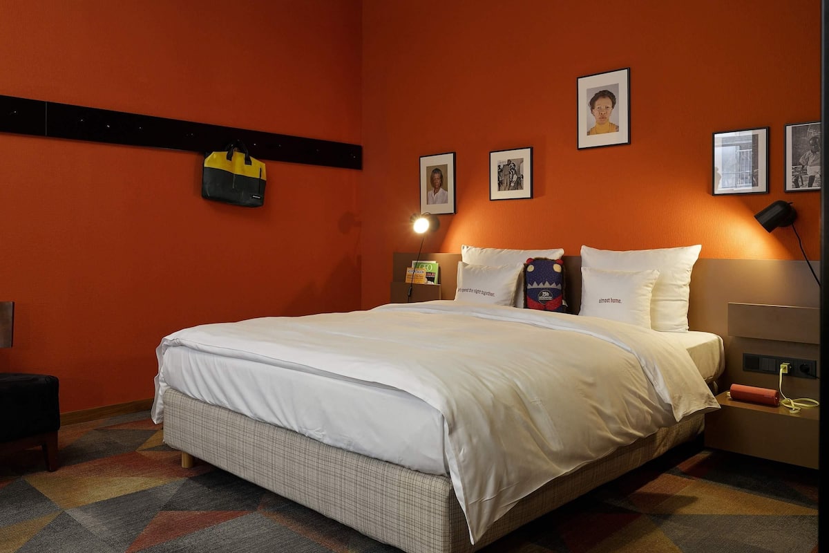 Quarto de hotel com uma cama grande, uma mesa de cabeceira, uma luminária e uma prateleira na parede com uma bolsa.