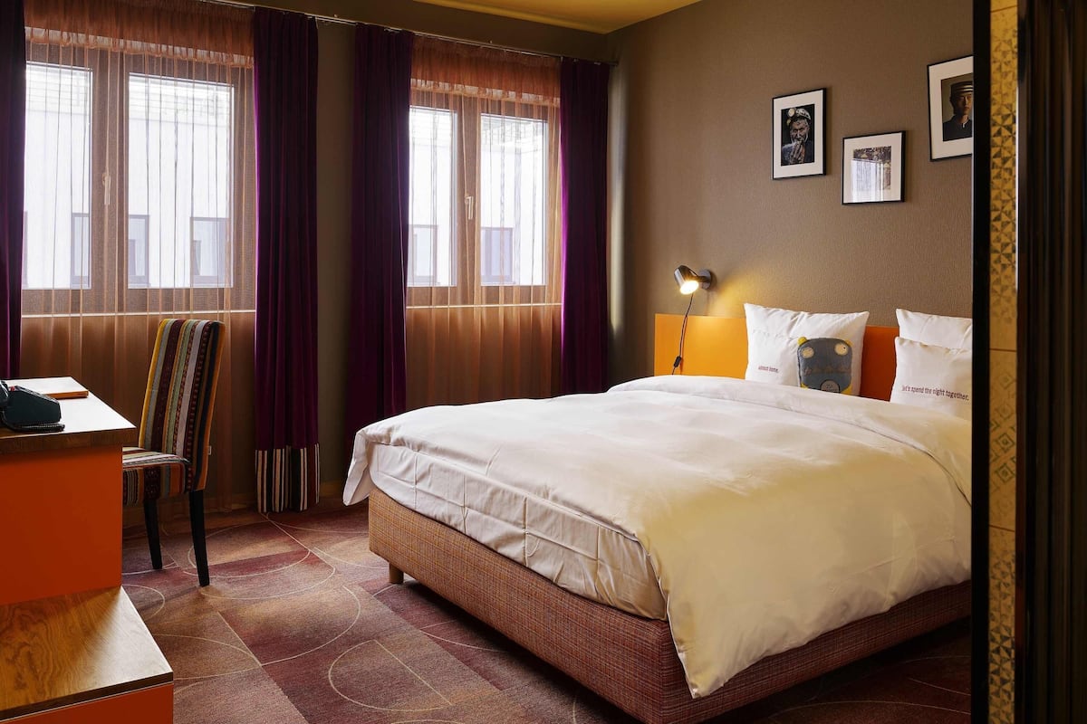 Quarto de hotel com uma cama grande, uma escrivaninha com cadeira e uma janela com cortinas.