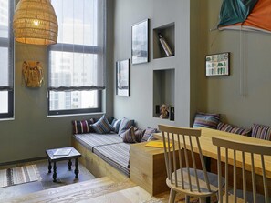 A cozy room with a wooden bench, a small table, and a chair. There are windows with sheer curtains, a hanging light fixture, and framed pictures on the wall. - 25hours Hotel The Trip (Frankfurt)