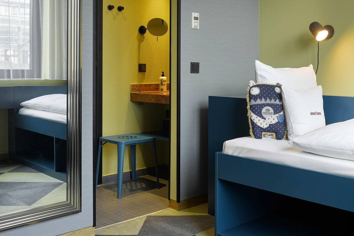 Quarto de hotel com duas camas, um banquinho pequeno, um espelho, uma pia e uma luminária fixada na parede.
