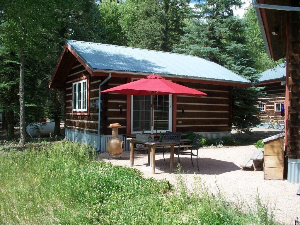 Une cabane rustique en rondins de bois, avec un parasol rouge, une table en bois et un banc.