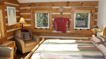 A log cabin room with a bed, a chair, a window with a view of trees, and a lamp.