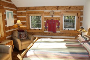 A log cabin room with a bed, a chair, a window with a view of trees, and a lamp.
